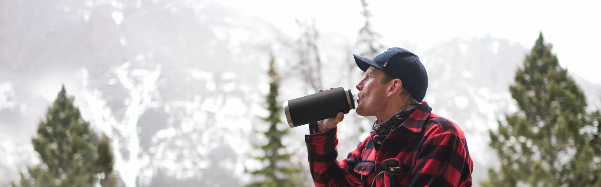 Person drinking from a black insulated bottle in a snowy forest setting