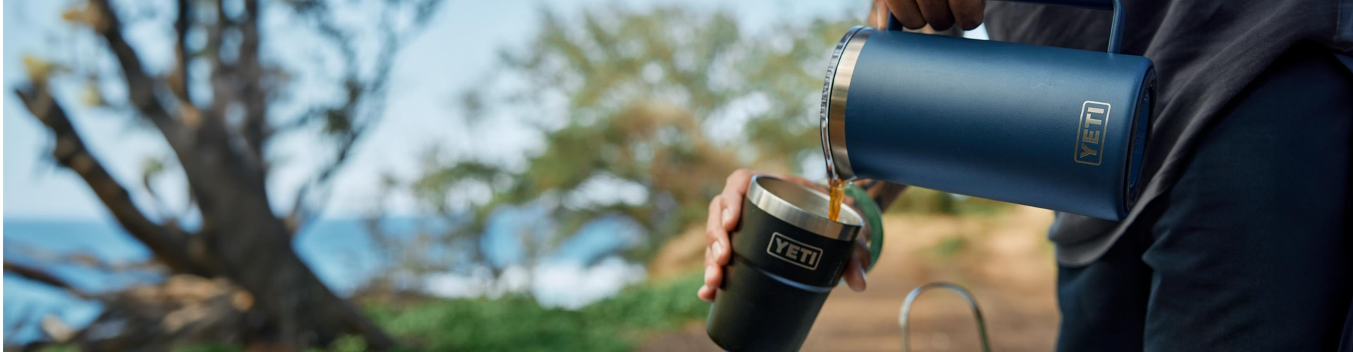 Person pouring coffee from a navy yeti french press into a black tumbler outdoors with a scenic background.