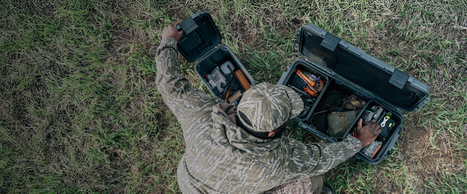 Person in camouflage gear with two black tool cases on a grassy field