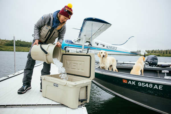 Person on a boat pouring ice into a Yeti cooler with two dogs nearby.