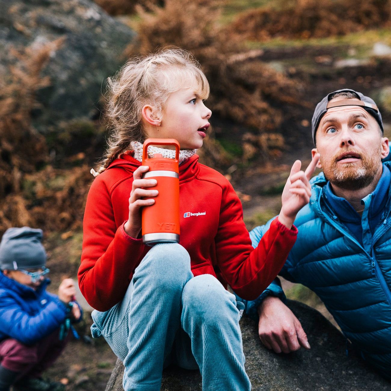 Man and two children sitting on rocks outdoors, with one child holding a red YETI bottle.