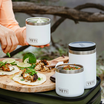 Three white YETI insulated tumblers with stainless steel rims are shown outdoors on a picnic-style setup. One tumbler is held by a person wearing an peach long-sleeve top, while two more tumblers sit on a green bag beside a wooden board with tacos, lime, and cilantro.