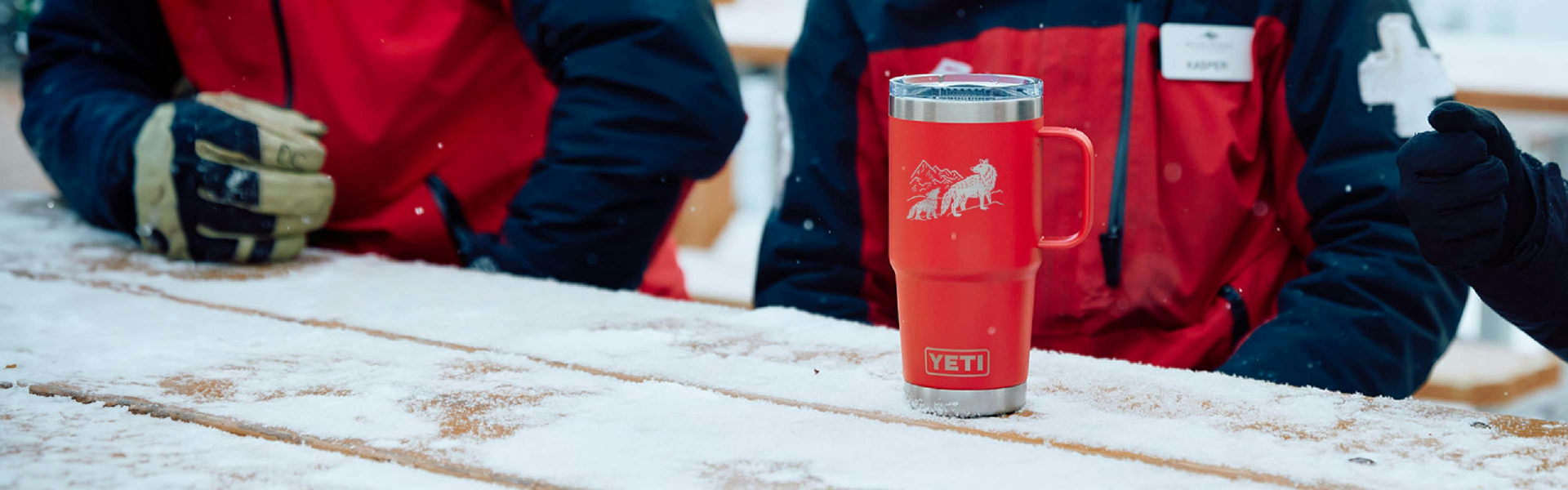 Red insulated mug on a wooden surface with people wearing red jackets in the background.