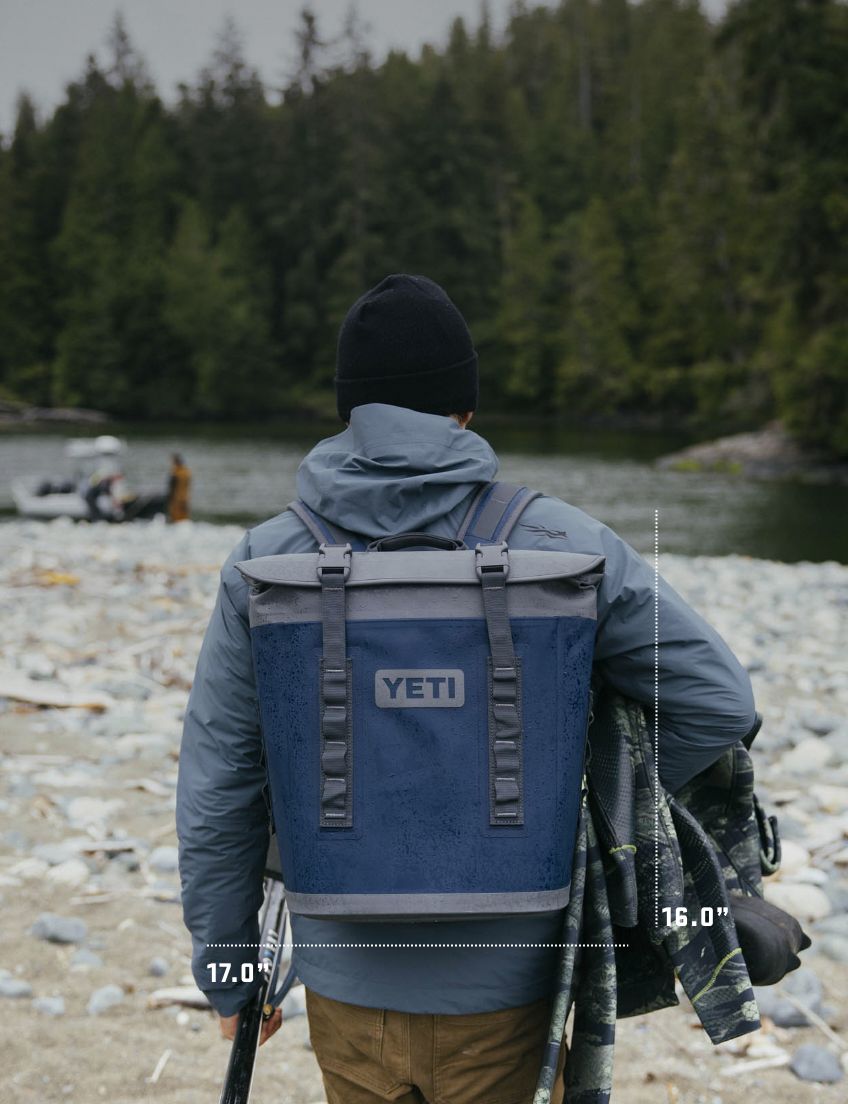Person with a YETI backpack standing by a river with trees in the background