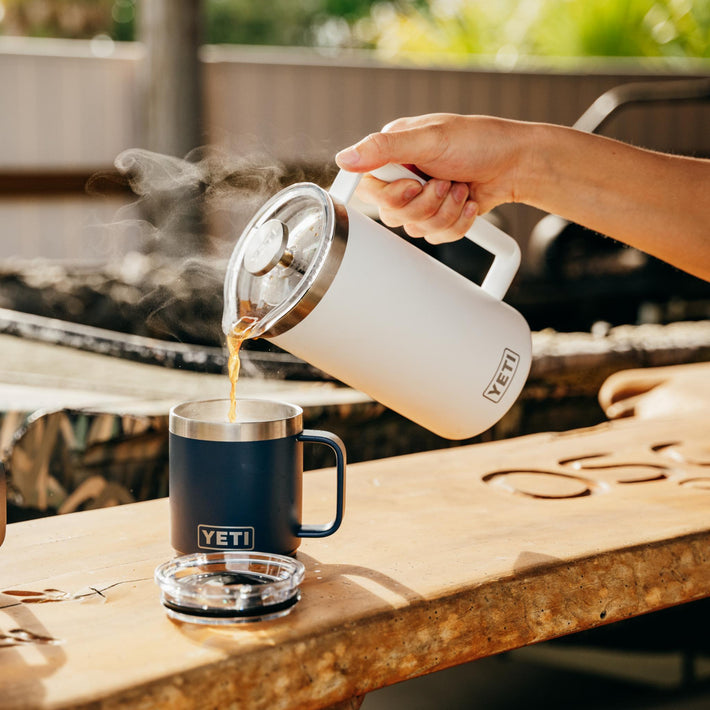 Person pouring coffee from a YETI french press into YETI mug on a wooden table outdoors.
