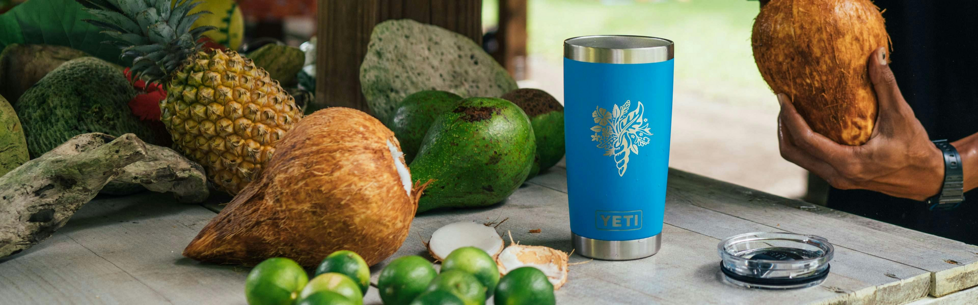Blue insulated tumbler with a tree design on a table with fruits and a person holding a coconut.
