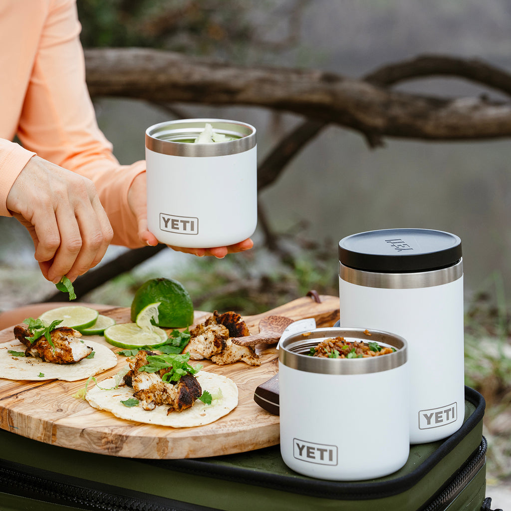 Three white YETI insulated food storage containers with stainless steel trim are shown outdoors on a picnic setup. One container is held by a person wearing a peach long-sleeve top, and two more sit on a green bag beside a wooden board with tacos, lime, and green onions.
