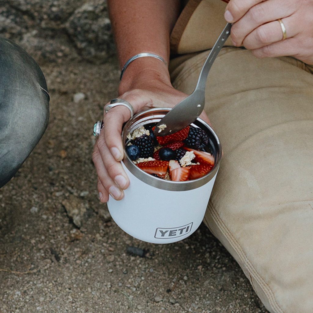 A close-up lifestyle shot of a person outdoors holding a white YETI insulated cup filled with fruit and granola, using a metal spoon to eat. The person is wearing khaki pants and silver jewellery.