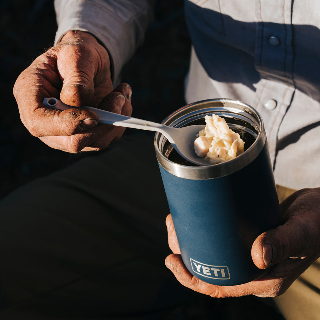 A close-up lifestyle shot of a person holding an open blue YETI insulated tumbler with a metal spoon, showing cream-coloured fat or butter inside. The YETI logo is clearly visible on the front of the tumbler.
