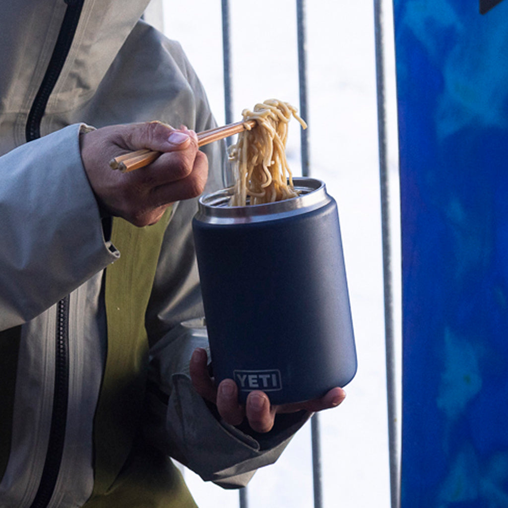 A person holding a YETI insulated can cooler in one hand and using chopsticks to lift noodles from the can with the other hand, shown in an outdoor setting.