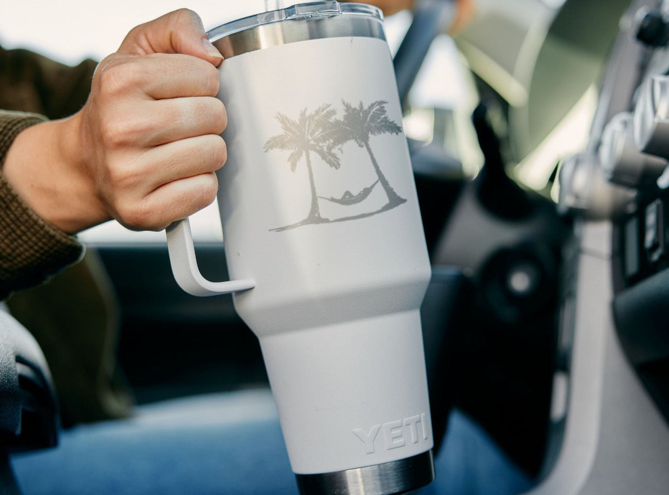 Person holding a white YETI mug with a palm tree design inside a car.
