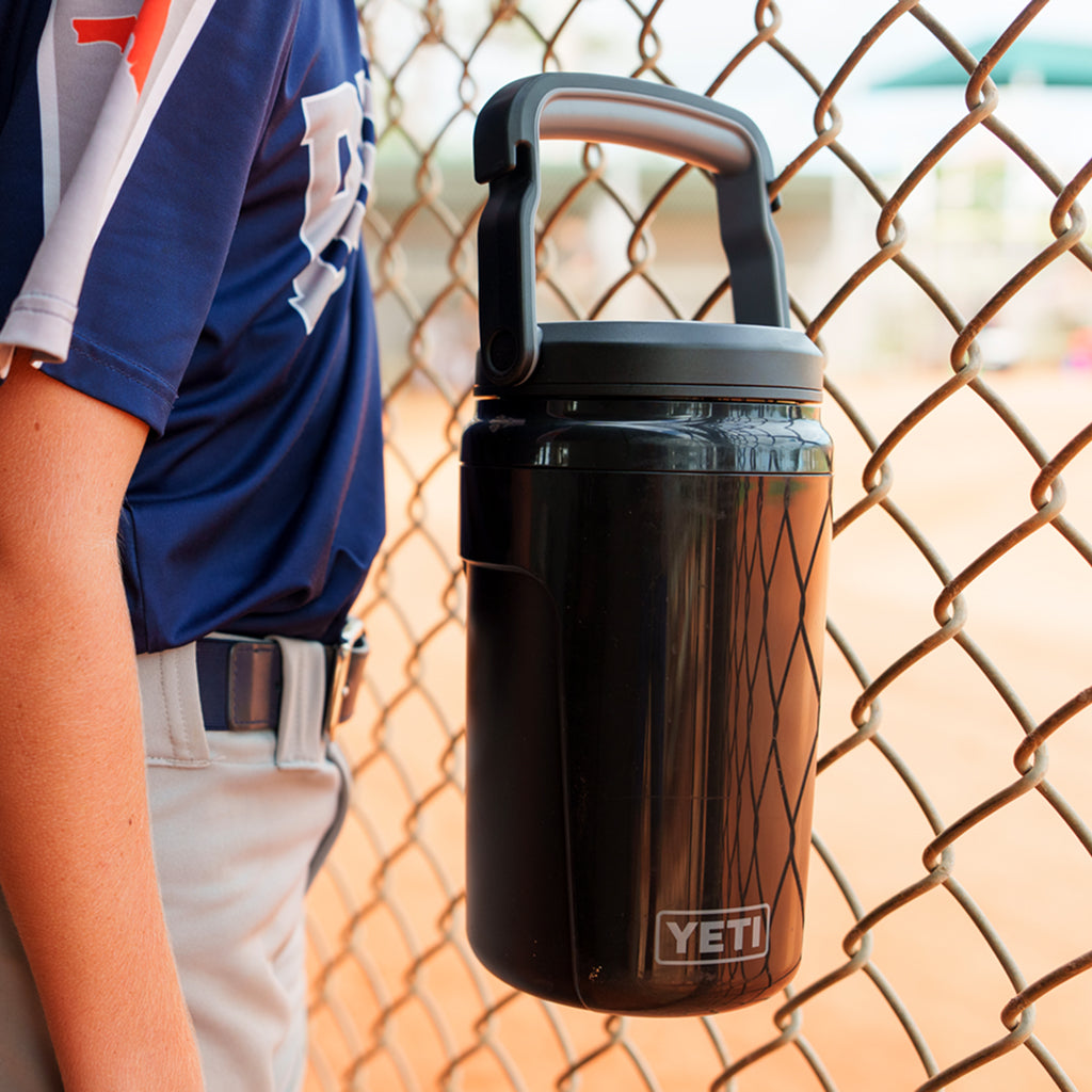 Person wearing a sports top holding a YETI Silo Jug with a chain-link fence in the background.