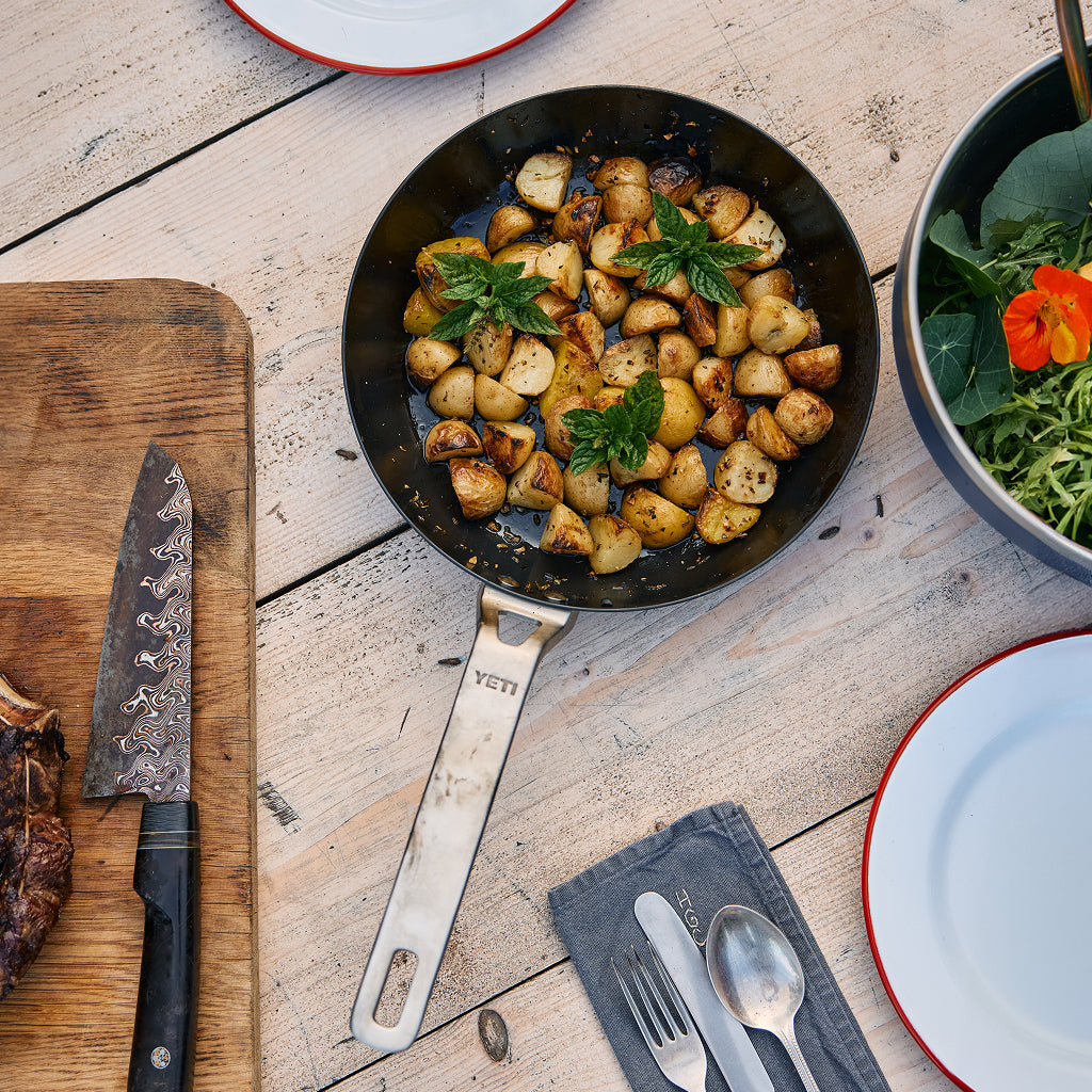 YETI frying pan with roasted potatoes on a wooden table with utensils and plates.