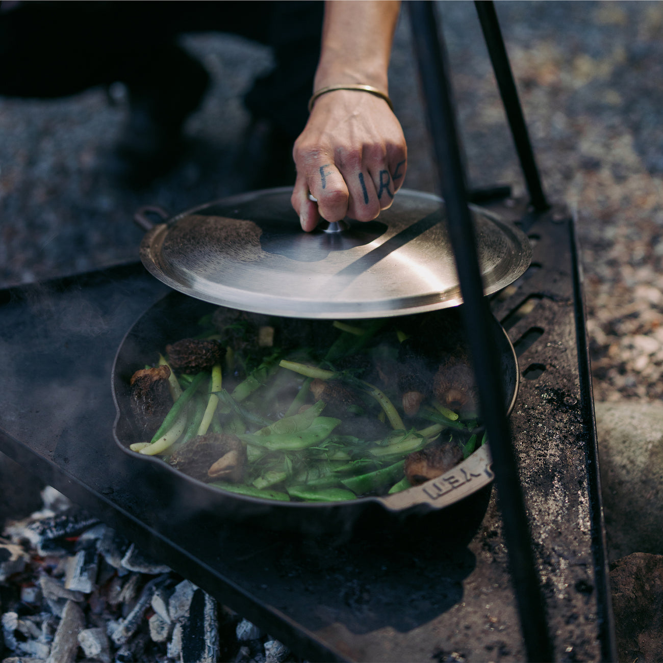 A cast iron skillet cooking vegetables over an open fire, with a hand lifting the lid. 