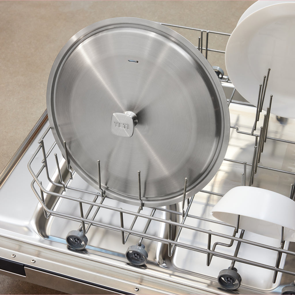 A stainless steel dishwasher rack holding a large round metal lid, with white dishes partially visible on the upper and lower racks.