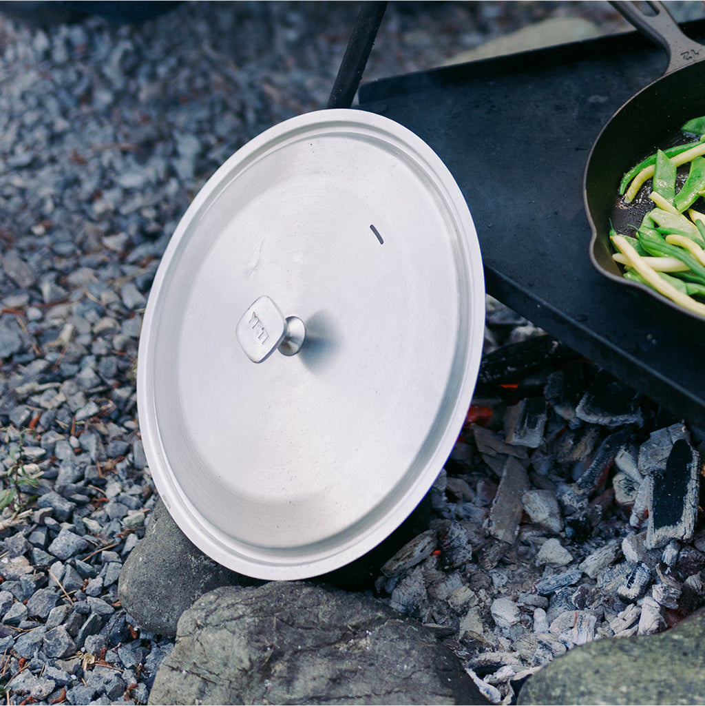 A silver metal pot lid with a small knob lies on rocks beside a black cast-iron skillet containing green beans over an outdoor fire.