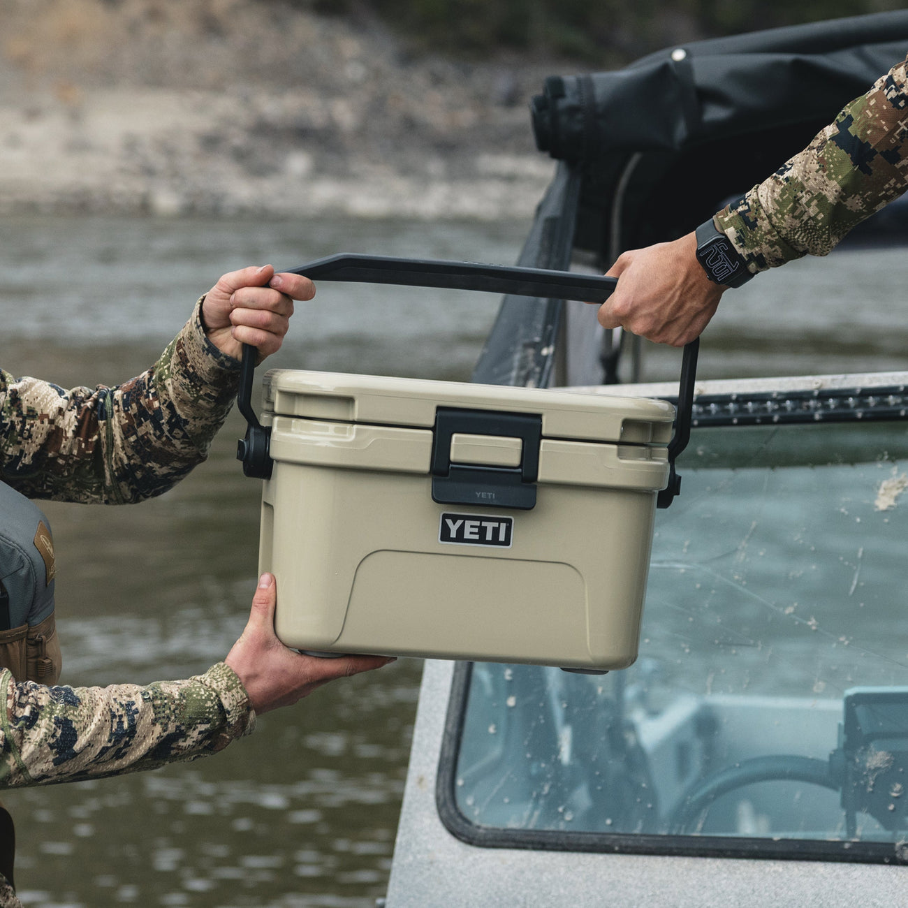 Two people lifting a YETI cooler out of a vehicle by a body of water.