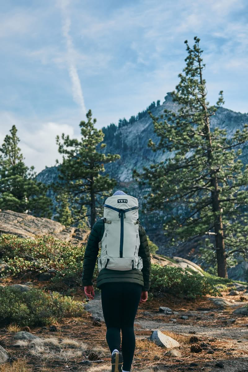 Person hiking with a backpack on a trail surrounded by trees and mountains.