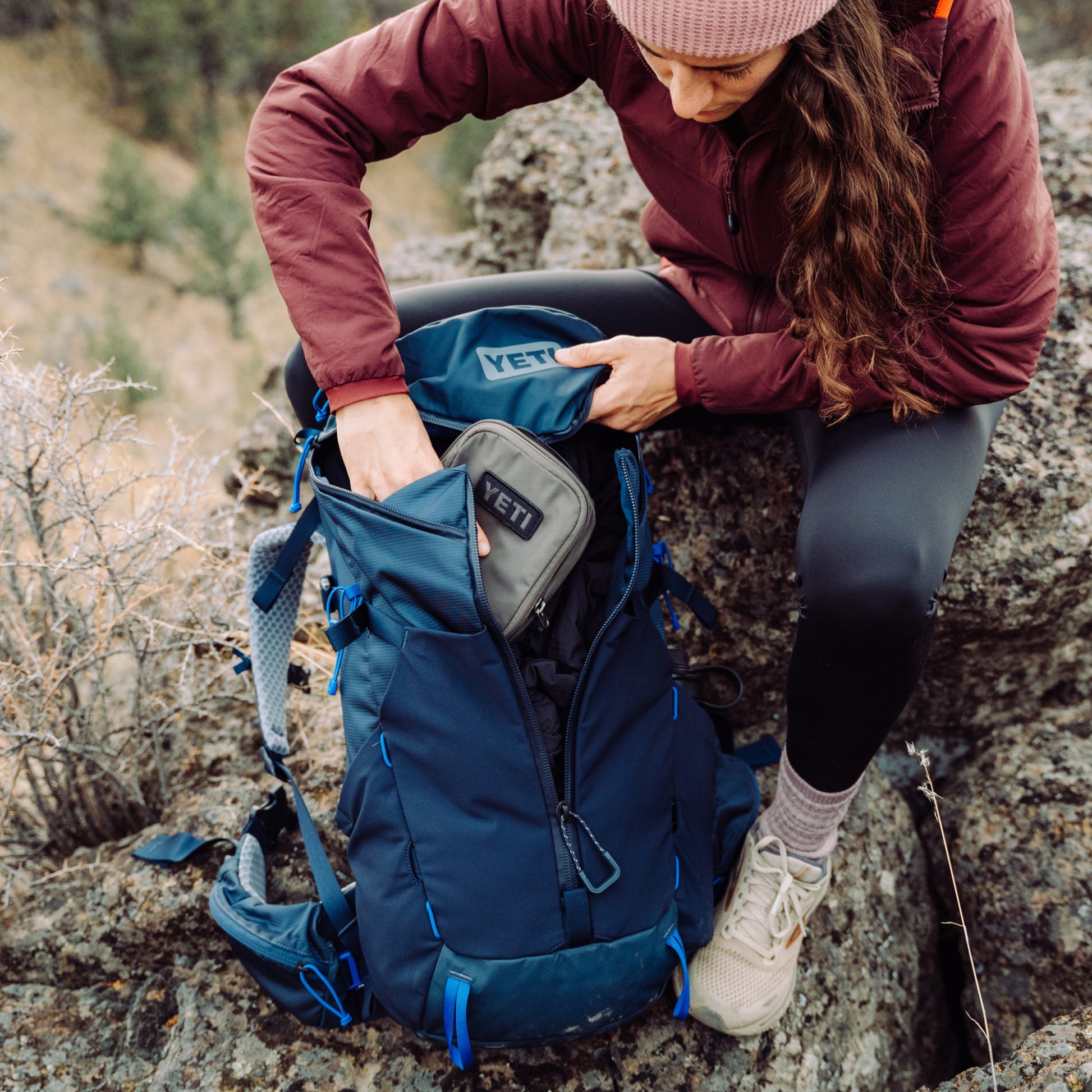 Person sitting on a rock with a blue backpack, opening it to reveal a Yeti packing cube.