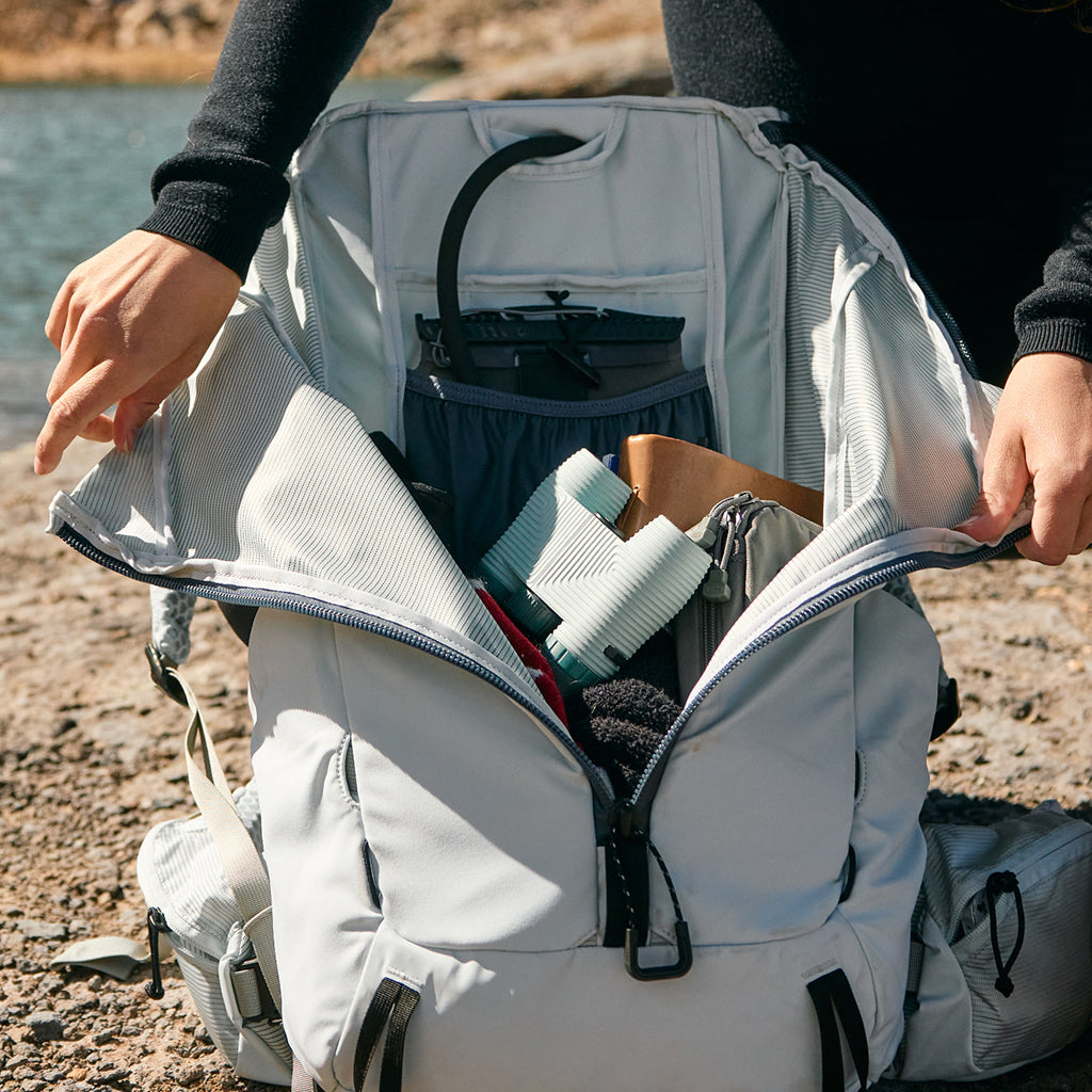 Person opening a backpack with items inside near a body of water.