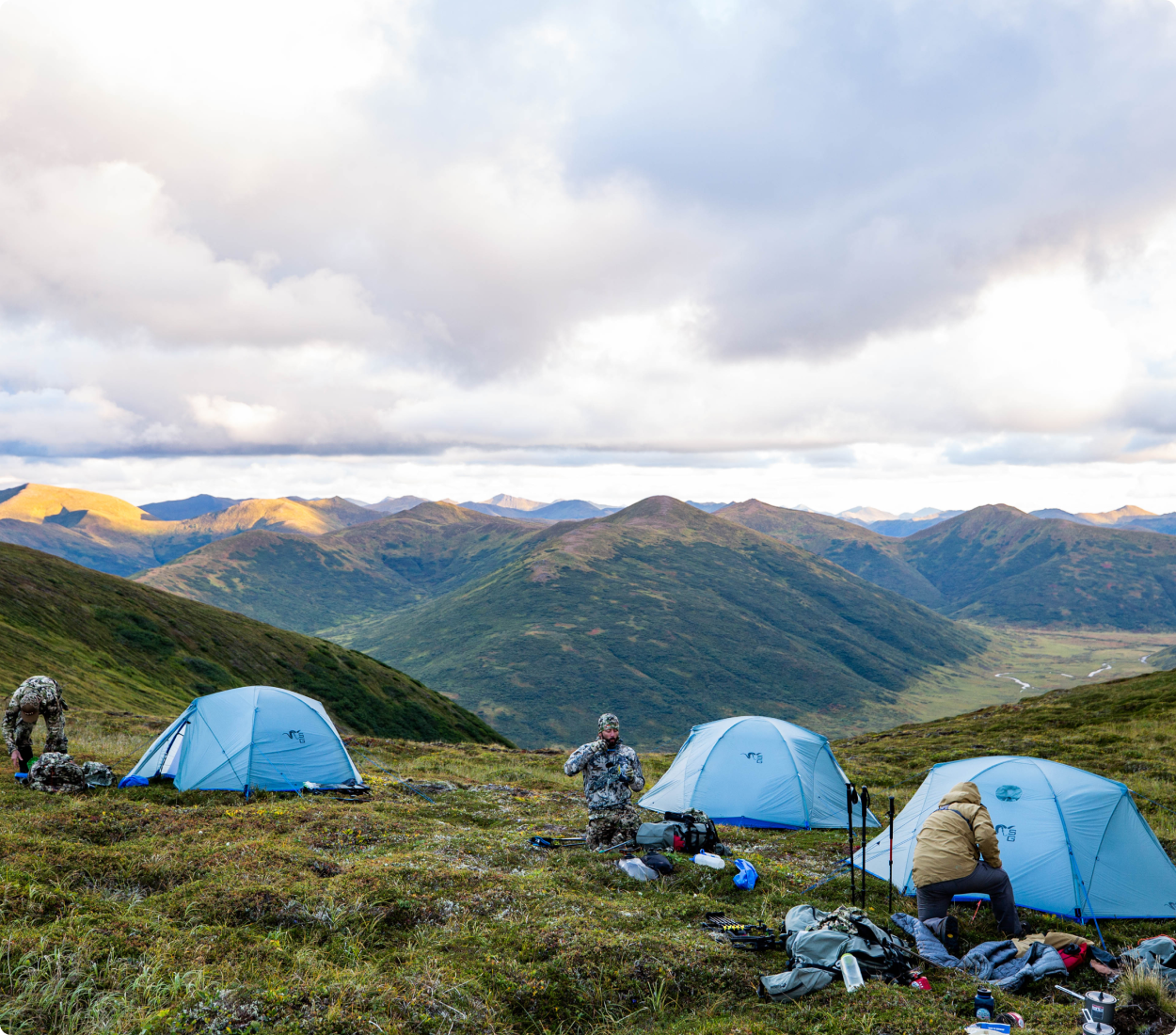 Camping scene with three tents and people in a mountainous landscape.