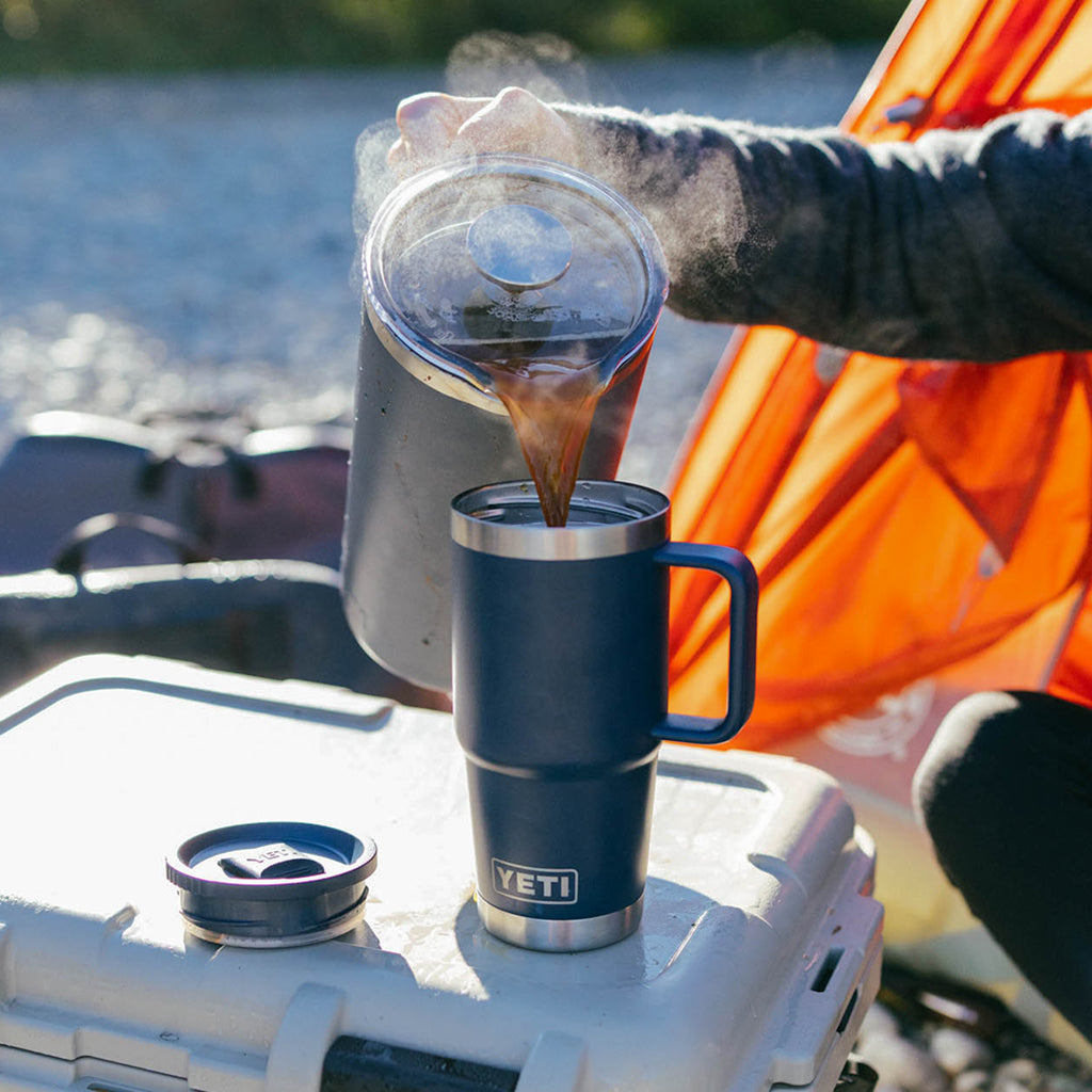 A person pouring hot coffee from a metal pot into a dark blue YETI insulated travel mug with a handle, set on top of a white cooler outdoors near water and an orange tent.