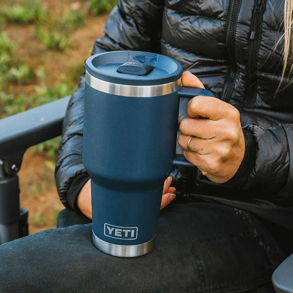 A navy blue YETI travel mug with stainless steel trim, held by a person wearing a black puffer jacket in an outdoor setting.