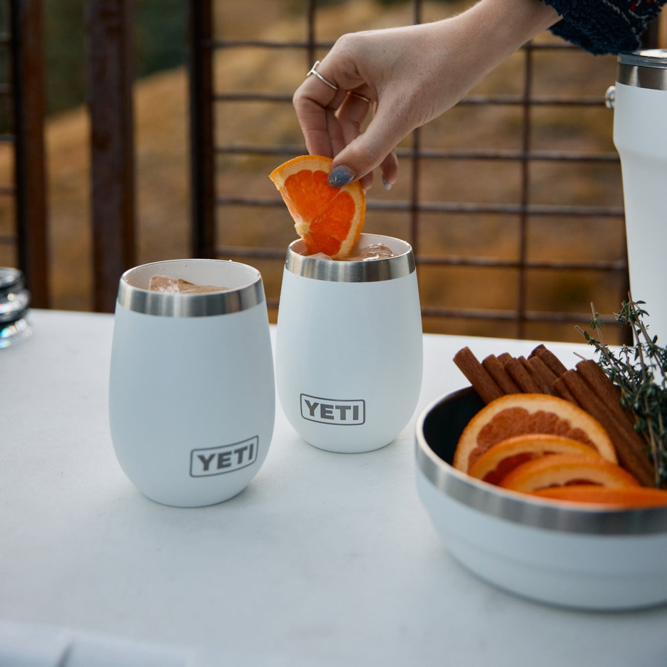 Two YETI wine tumblers with a person adding an orange slice, set on a table outdoors.