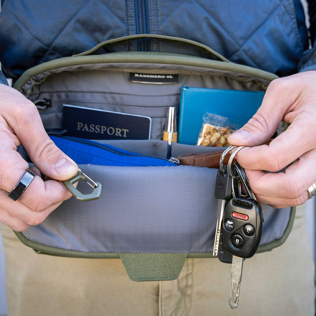 An olive green and gray sling bag worn across a person's waist, shown open with hands holding out accessories including a car key fob, ring, and a blue zip pouch. 