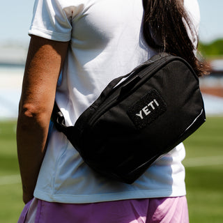 A person outdoors wearing a black YETI crossbody bag across their torso, with a white shirt and pink skirt visible against a blurred green lawn background.