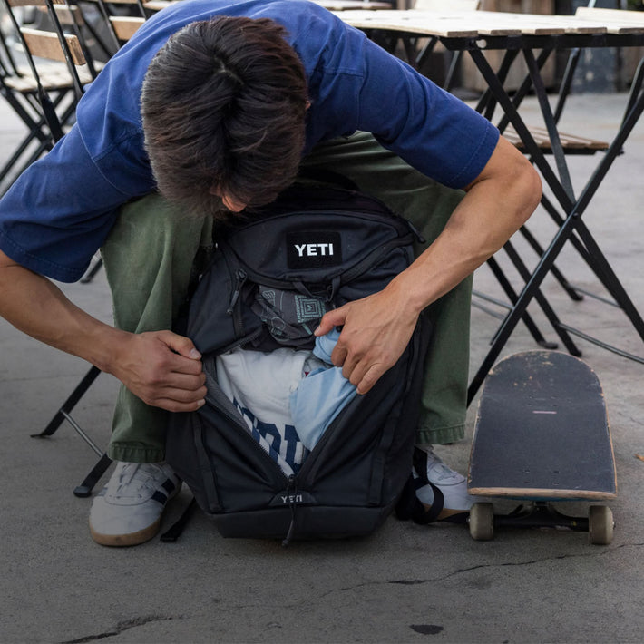 A person crouches beside a black YETI backpack, opening the front pocket to put or retrieve items, with a skateboard nearby on an outdoor patio.