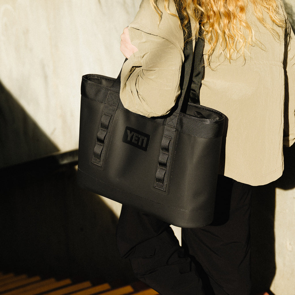 Person holding a black YETI Camino Zip tote bag against a neutral background