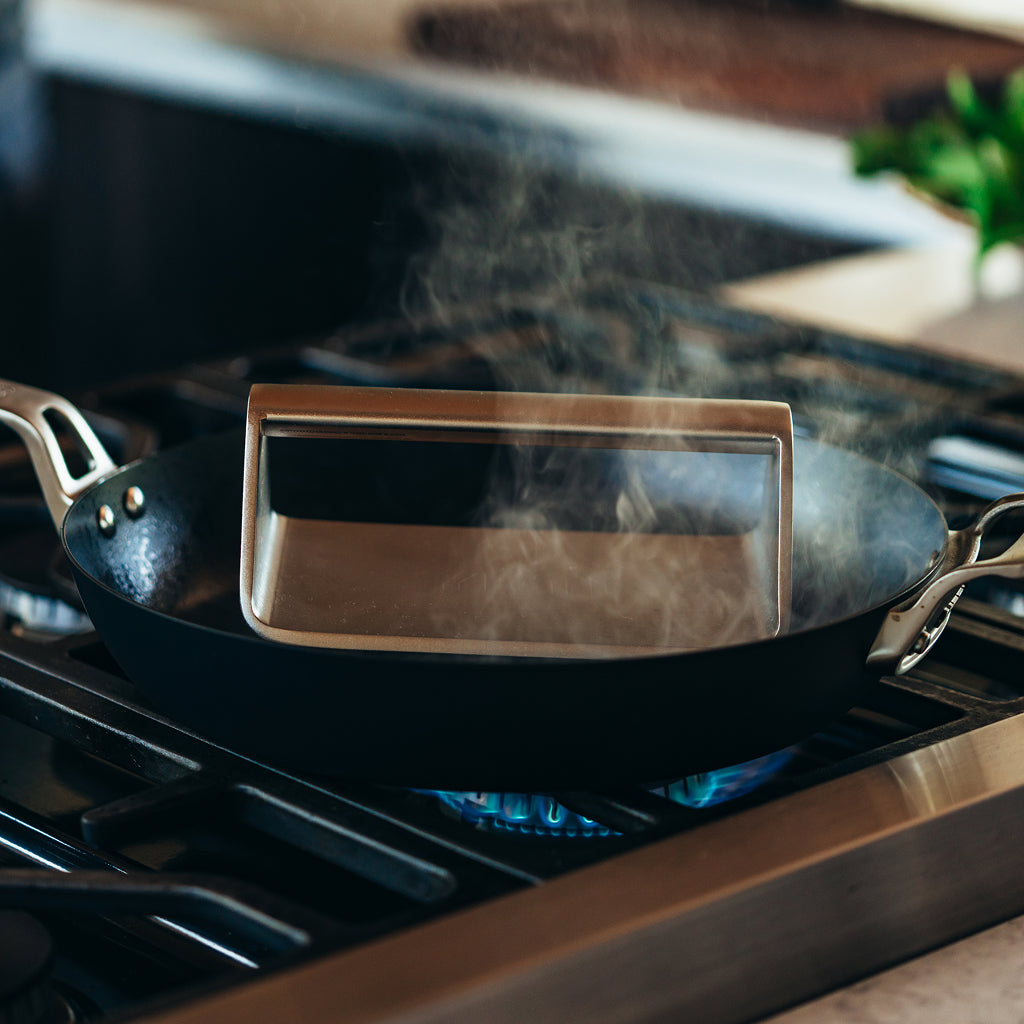 A YETI grill press resting inside a black frying pan on a gas stovetop with visible blue flame and steam rising.