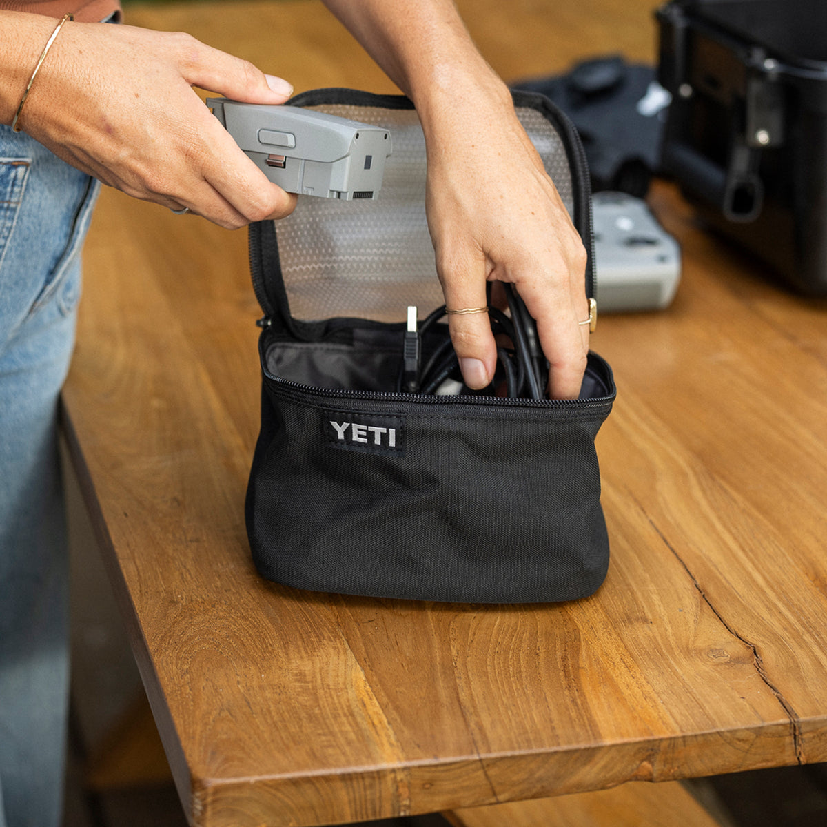 A person's hands placing a camera into a black YETI zippered carrying case on a wooden table, with other camera gear blurred in the background.