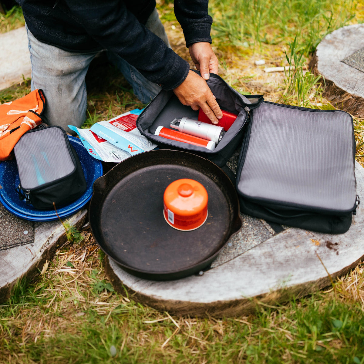 A person outdoors organizes camping gear in a black zippered pouch beside a black cast-iron skillet with an orange fuel canister, along with other camping items on a stone slab in grass.
