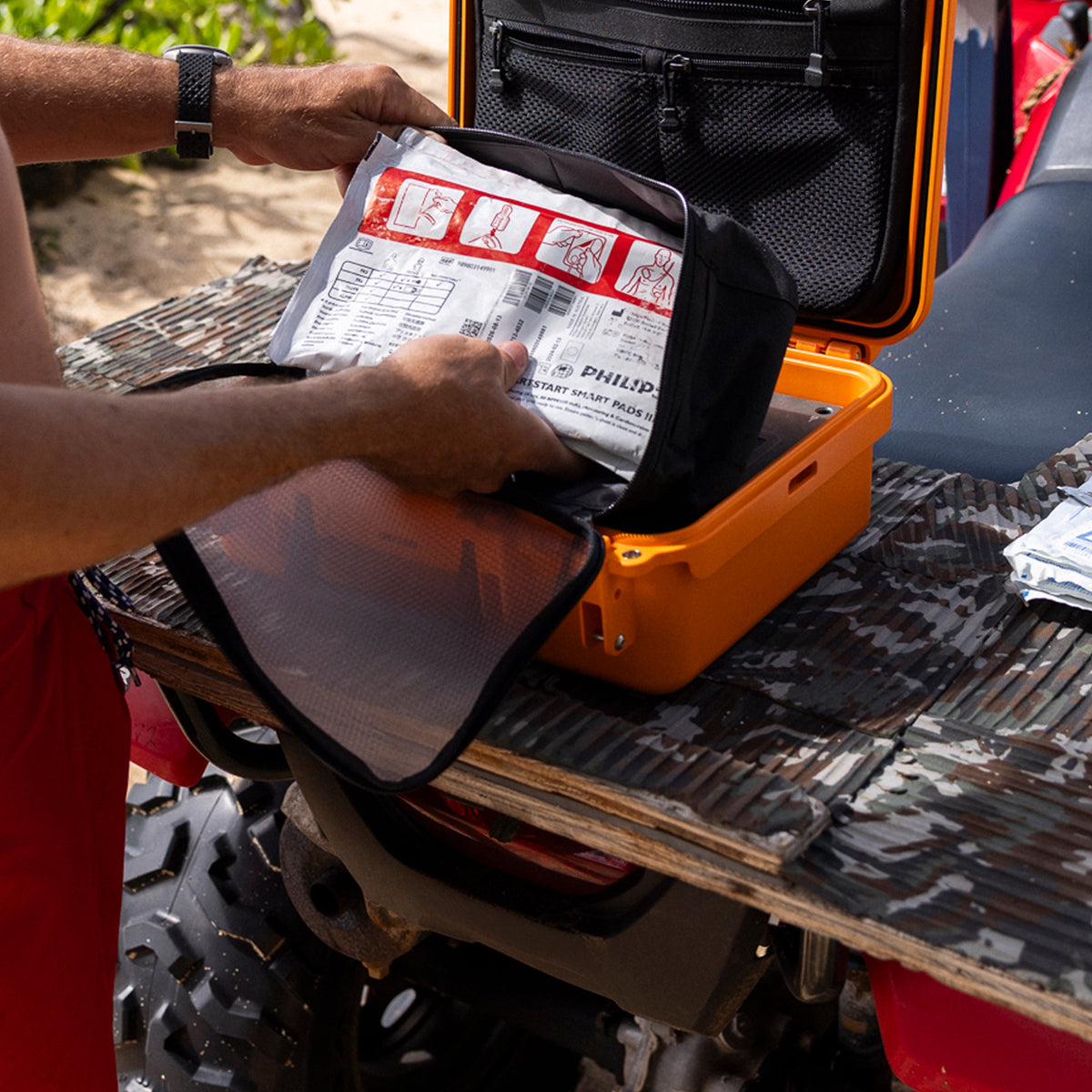 A person's hands are shown placing a white and red medical instruction packet into an orange hard case with a black mesh organizer, set on the back of a vehicle.