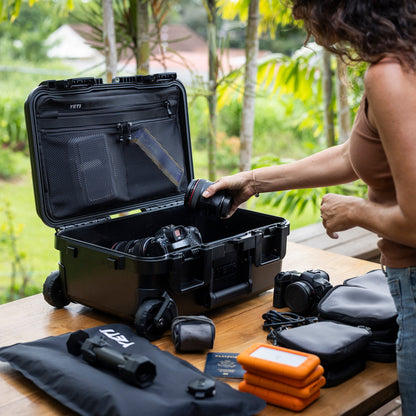 A woman organizes camera gear into an open black Yeti hard case on a wooden table, with outdoor greenery in the background. The case is shown with cameras, lenses, pouches, and an orange hard drive encloser.