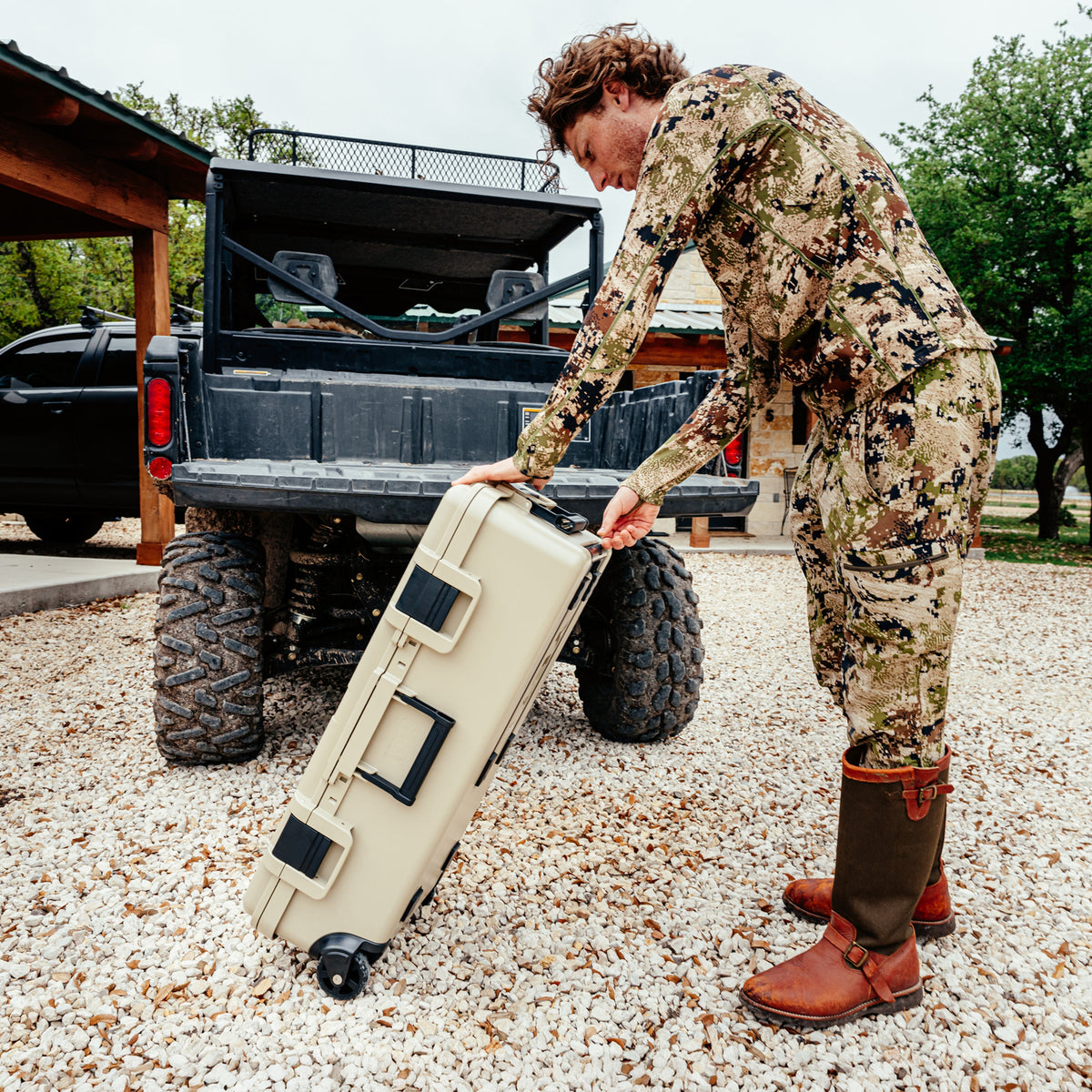 A man in a desert camouflage hunting suit and brown boots pushes a beige hard-shell rolling case on a gravel driveway next to a black off-road truck.
