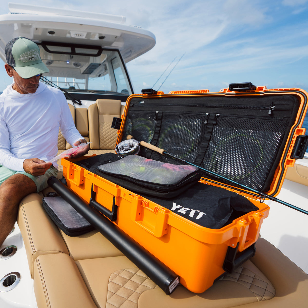 A man sitting on a boat beside an open bright orange YETI hard case filled with fishing gear, including a black rod case and tackle bags, under a blue sky.