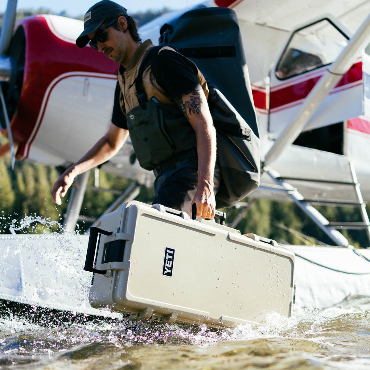 A man in outdoor gear carries a large tan-coloured YETI hard case through splashing water next to a red and white seaplane.