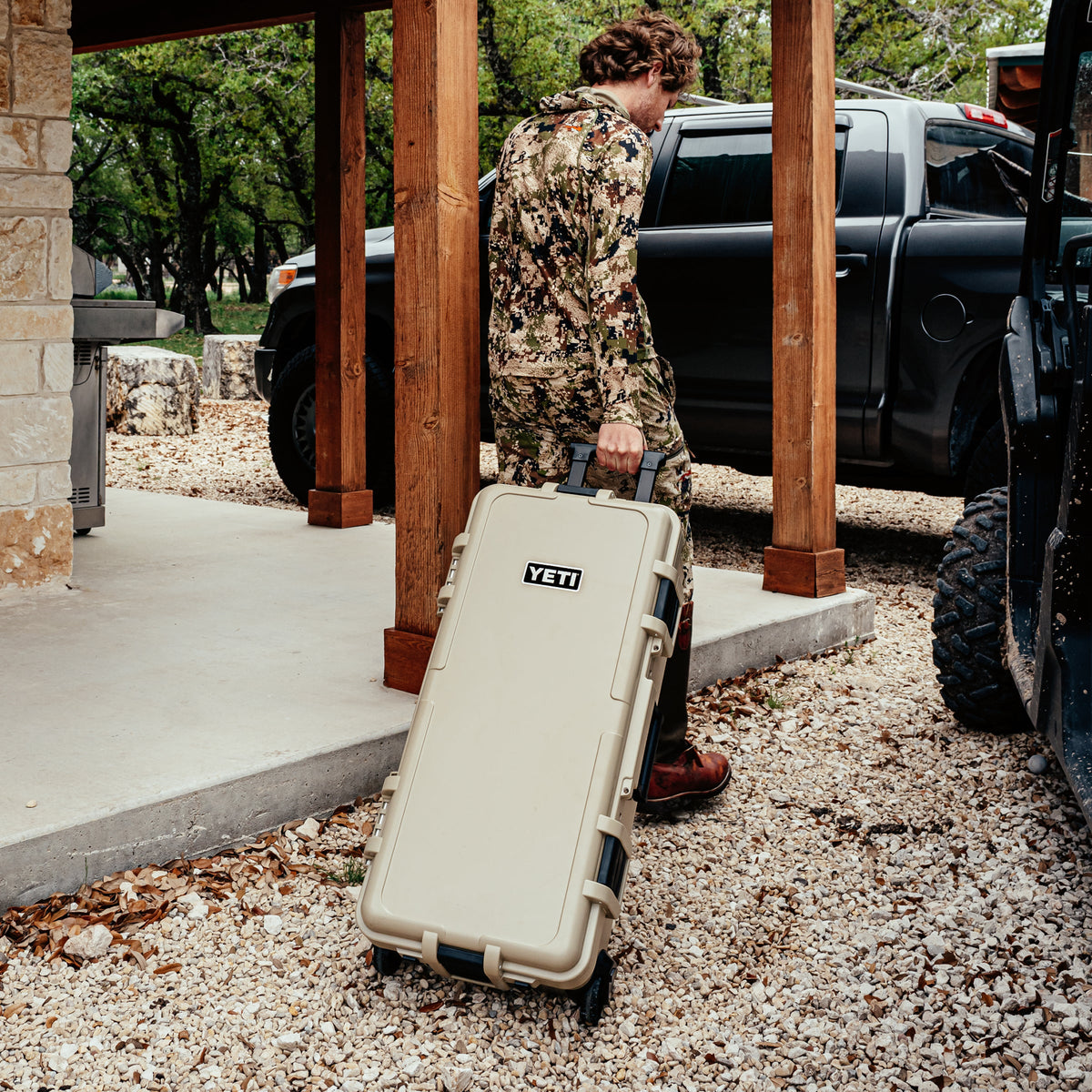 A person in a camouflage jacket carries a beige YETI rolling hard case on a gravel porch next to a black pickup truck.