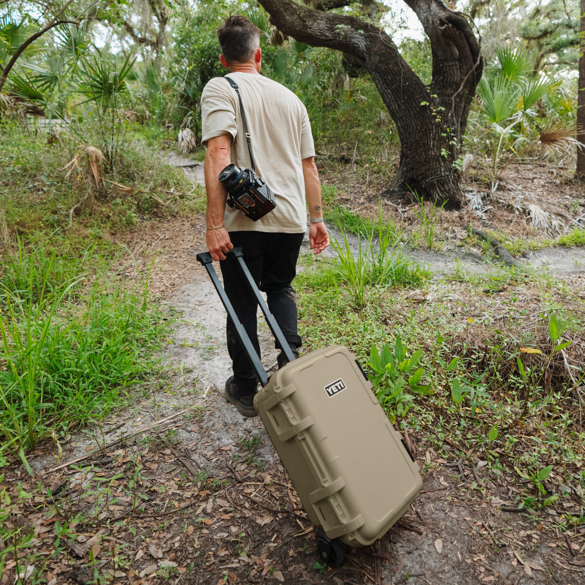 A man walking on a forest path pulling a tan hardshell rolling scase with a camera over his shoulder.