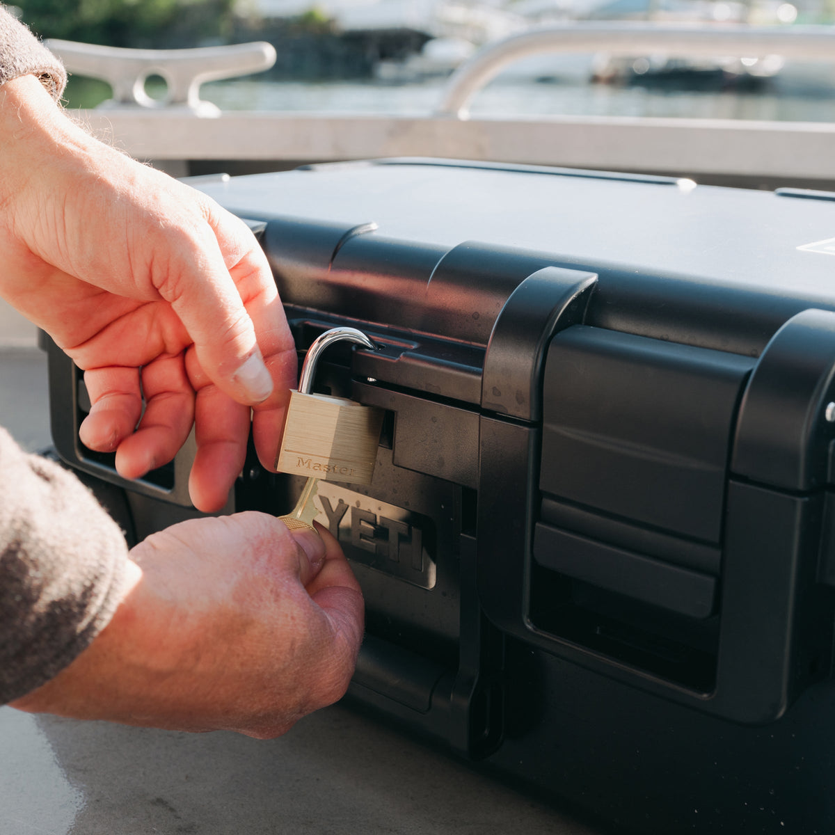 A close-up of a person's hands securing a black hard case with a brass padlock on a boat deck, with water and boat railings blurred in the background.