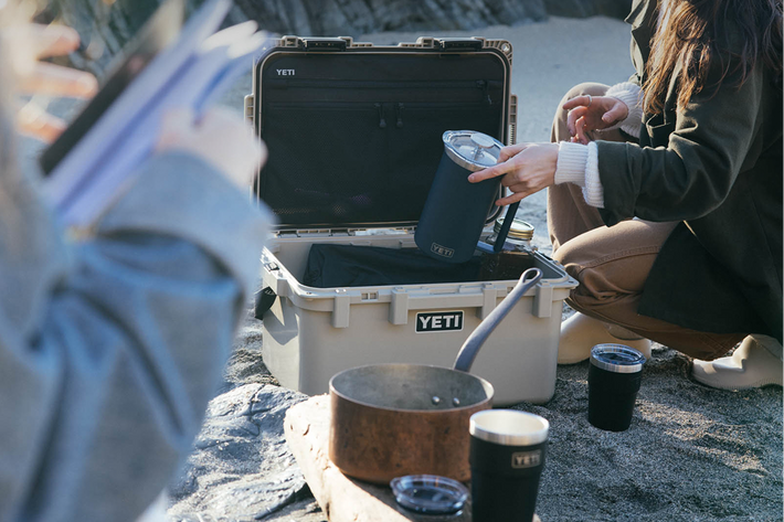 Person preparing a meal outdoors with a YETI box on a rocky beach.