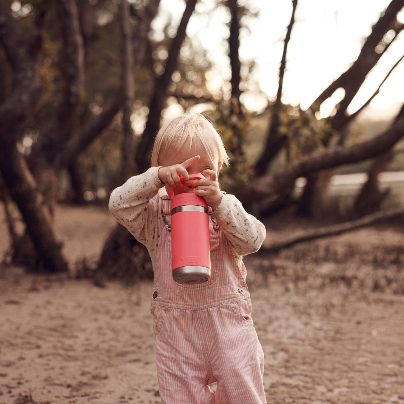 Child holding a pink YETI bottle in a natural setting with trees and water.