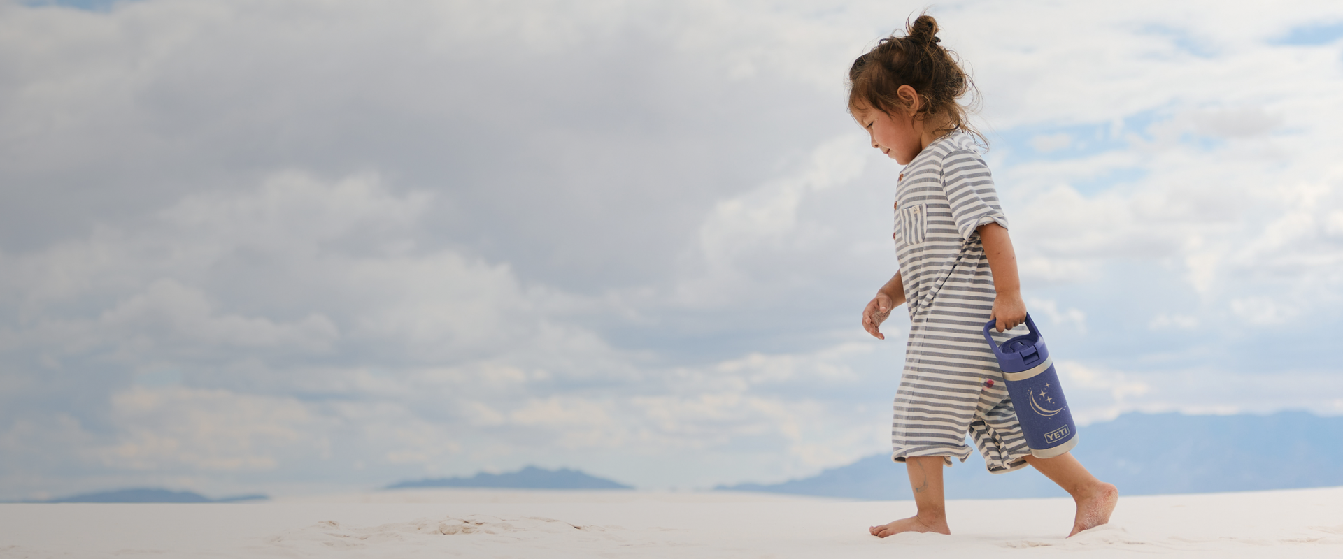 Child in a striped dress walking on a white sand dune with a purple bottle.