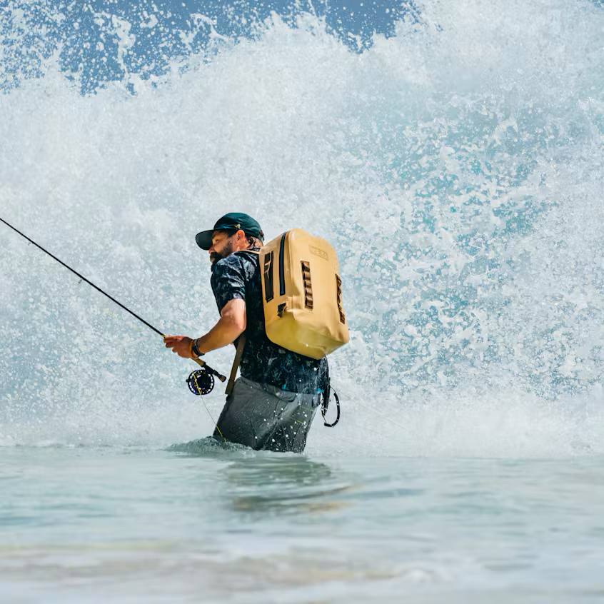 A man in a fishing wetsuit stands in ocean water holding a rod, with a large breaking wave behind him and a tan backpack mounted on his back. The backpack has visible vertical branding that reads 'YETI'.