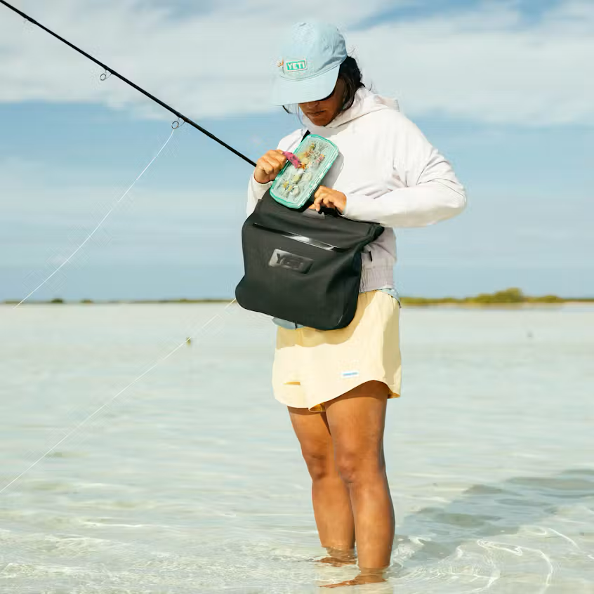 Person fishing in shallow water wearing a cap and light clothing, with a black dry bag.