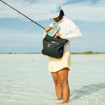 Person fishing in shallow water wearing a cap and light clothing, with a black dry bag.