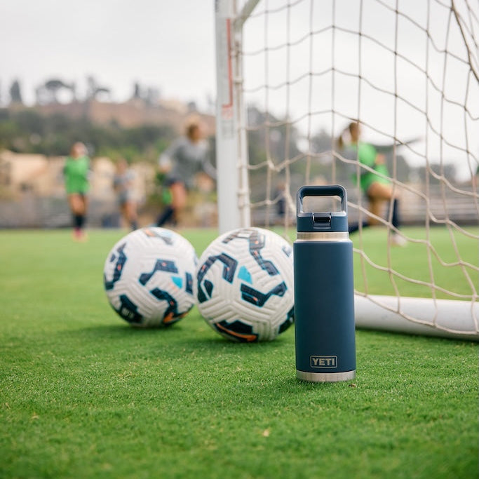 Two footballs and a blue YETI water bottle on a grass field with a goalpost in the background.